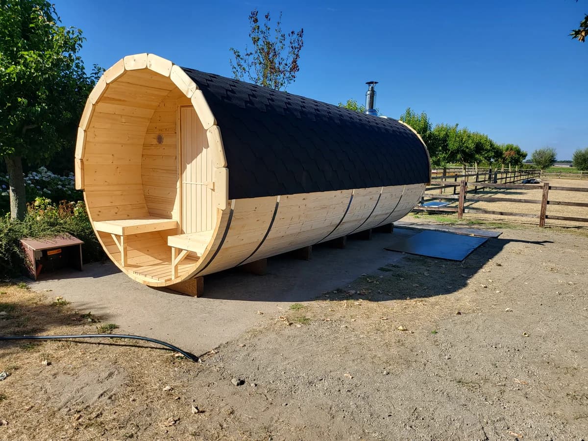 A barrel sauna outdoors: open terrace with two benches, black shingle roof and a chimney
