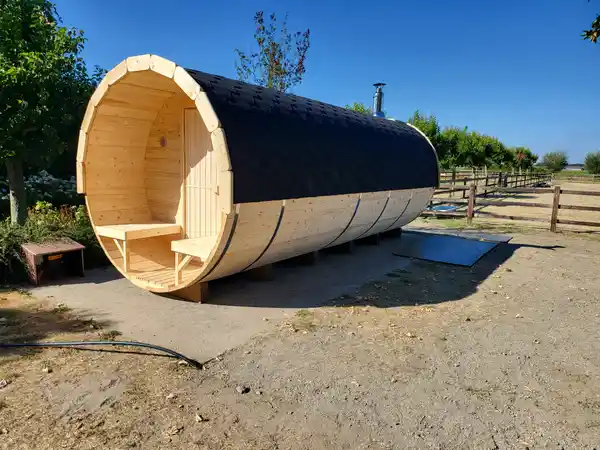 A barrel sauna outdoors: open terrace with two benches, black shingle roof and a chimney